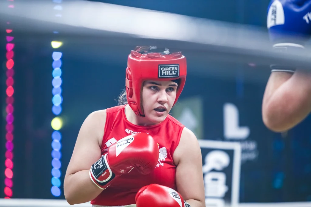 Agata Kaczmarska Kobieta w czerwonym stroju i kasku bokserskim podczas walki na ringu, skupiona i gotowa do ataku, w tle kolorowe światła i fragment sylwetki przeciwniczki.