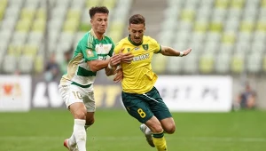 Dwóch piłkarzy w trakcie dynamicznej walki o piłkę na boisku podczas meczu, jeden ubrany w zielono-biały strój, drugi w żółto-zielony, w tle trybuny stadionu.