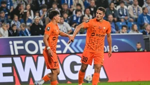 Dwóch piłkarzy w pomarańczowych strojach drużyny KGHM Zagłębie Lubin stoi na murawie stadionu, za nimi widoczna publiczność i reklamy na bandach.