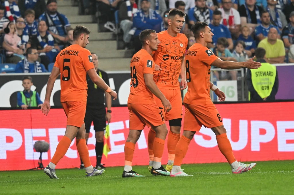 Zagłębie Lubin Grupa piłkarzy drużyny ubraną w pomarańczowe stroje rozmawia i gestykuluje na boisku podczas meczu, w tle widać tłum kibiców oraz banery reklamowe przy trybunach.