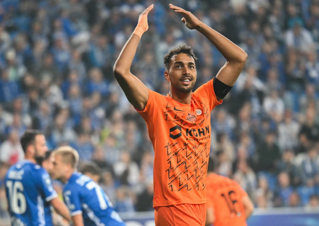 Lech Poznań - Zagłębie Lubin, 12.09.2025 r. Piłkarz w pomarańczowym stroju unosi ręce w geście radości na tle stadionu pełnego kibiców, w tle widoczni zawodnicy drużyny przeciwnej w niebieskich koszulkach.