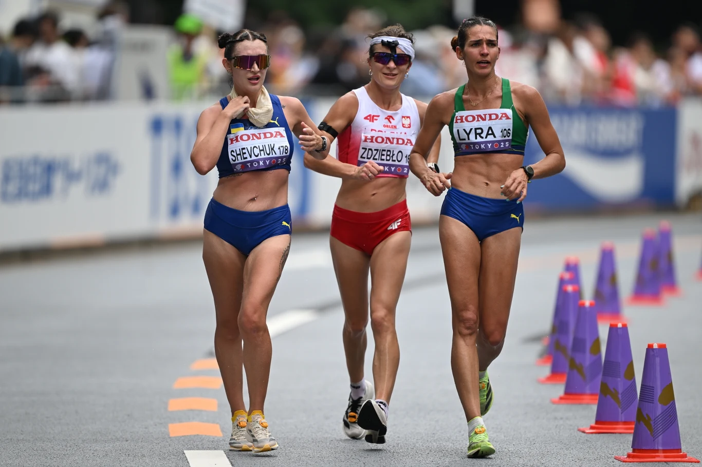 Trzy zawodniczki w sportowych strojach idą razem trasą wyścigu, otoczone elementami organizacyjnymi takimi jak pachołki oraz widoczne identyfikatory na ubraniach.