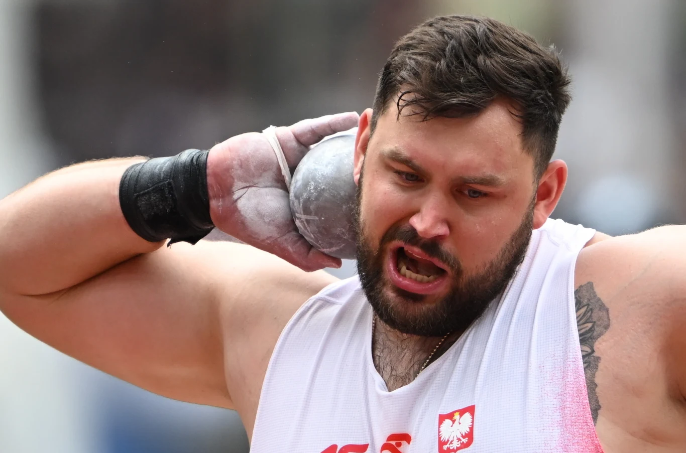 Mężczyzna podczas wykonywania rzutu kulą, skupiony wyraz twarzy, na ręce widoczna rękawica ochronna oraz tatuaż, na białej koszulce godło Polski.