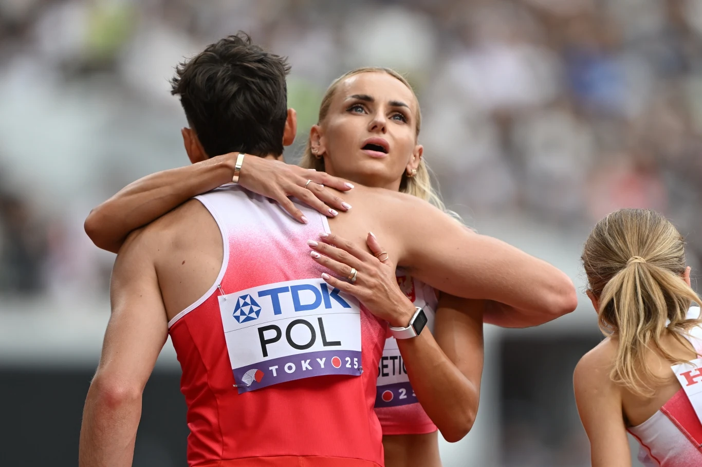 Dwóch sportowców w biało-czerwonych strojach reprezentacji Polski, przytulających się po zakończonym biegu, na plecach widoczny napis POL i plakietka Tokyo 2020, w tle rozmazane sylwetki publiczności.