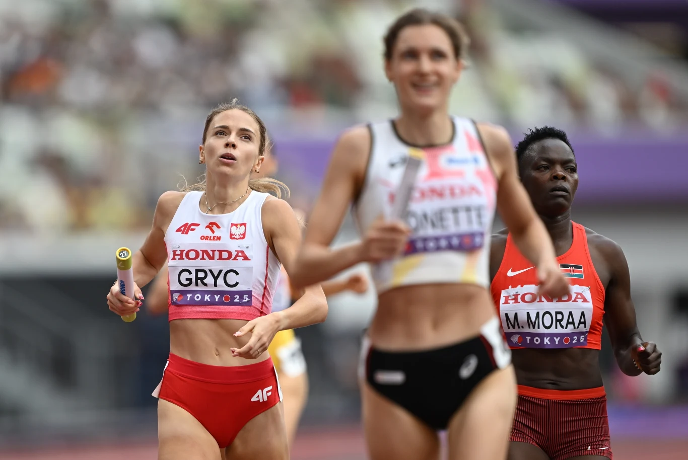 Zawodniczki biegnące w sztafecie lekkoatletycznej na stadionie podczas zawodów olimpijskich, każda w stroju reprezentacji swojego kraju, widoczna koncentracja i wysiłek sportowy.