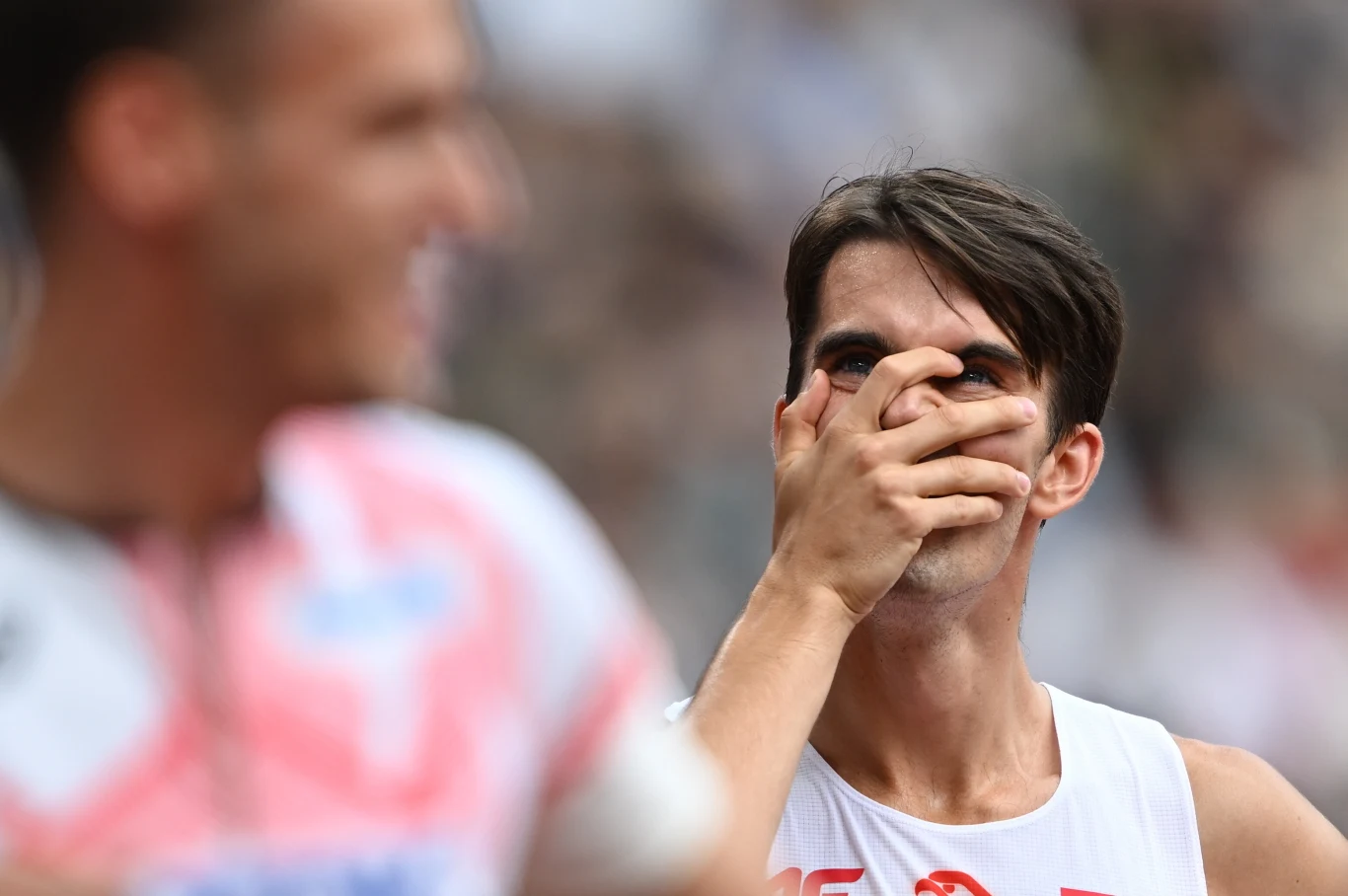 Sportowiec w białym stroju z numerem zakrywa dłonią usta, patrząc w przód z wyrazem skupienia na twarzy, a na pierwszym planie widoczny jest rozmazany profil innego sportowca, co sugeruje moment emocji podczas zawodów sportowych.