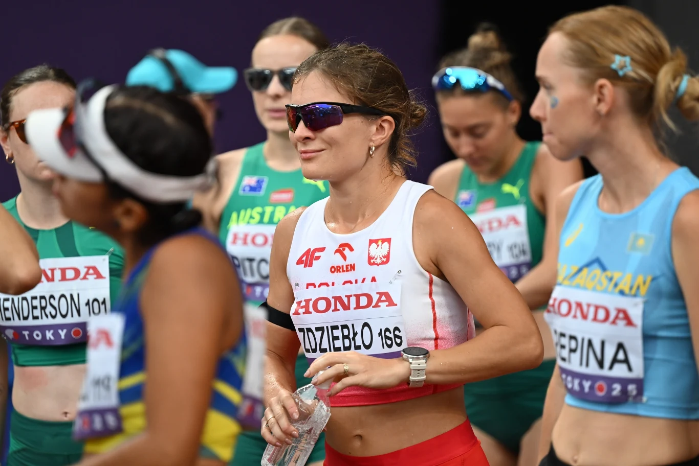 Zawodniczka reprezentująca Polskę ubrana w strój sportowy, przygotowuje się do startu podczas zawodów lekkoatletycznych w otoczeniu innych lekkoatletek z różnych krajów.