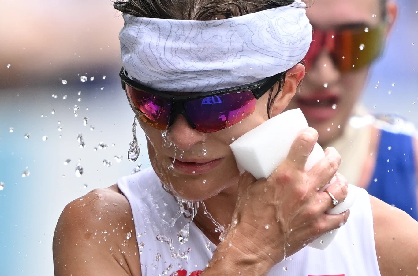 Katarzyna Zdziebło Zbliżenie na sportowca z opaską na głowie i okularami przeciwsłonecznymi polewającego się wodą z białej gąbki, na twarzy widoczne krople wody odbijające światło.