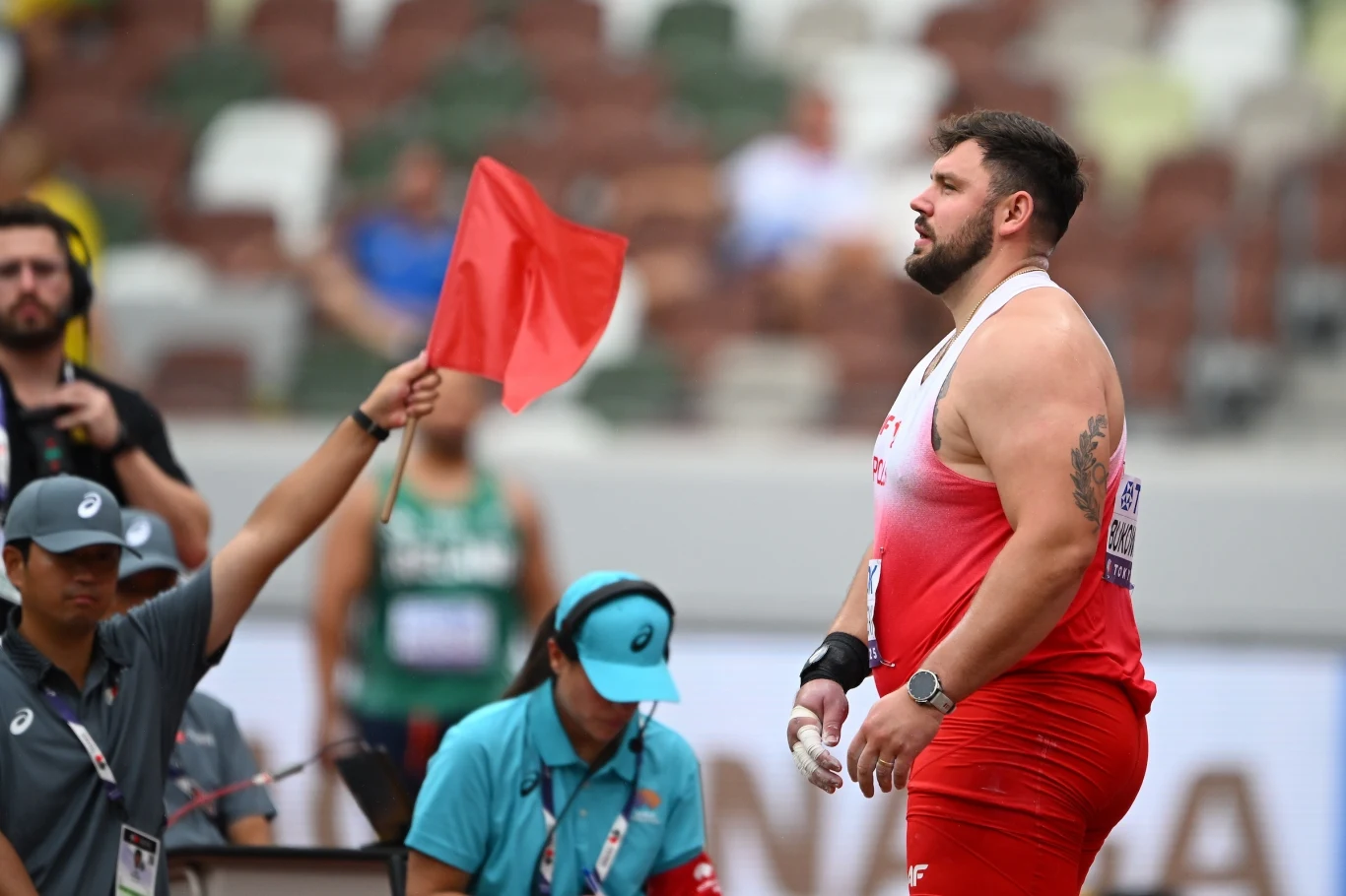 Zawodnik w czerwonym stroju stoi na stadionie lekkoatletycznym, w tle sędzia podnosi czerwony chorągiewkę sygnalizującą faul podczas konkurencji pchnięcia kulą.