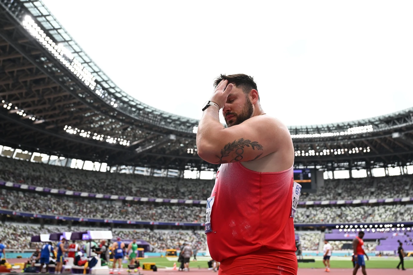 Lekkoatleta w czerwonym stroju sportowym z widocznym tatuażem na ramieniu stoi na stadionie, sprawia wrażenie zamyślonego lub zamartwionego, trzyma rękę na głowie, w tle widać innych sportowców oraz trybuny pod zadaszeniem.