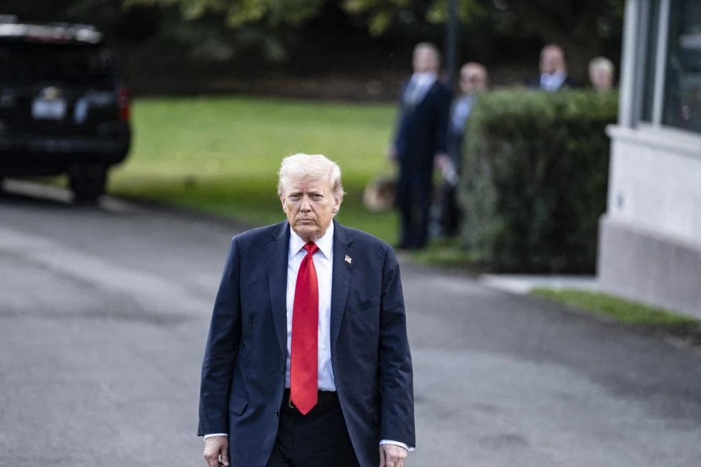 Prezydent USA Donald Trump Mężczyzna w garniturze z czerwoną krawatą idzie chodnikiem na tle rozmytego, zielonego terenu, w tle kilku mężczyzn w garniturach.