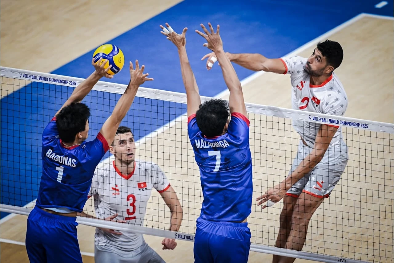 Mecz Filipin z Tunezją w meczu otwarcia siatkarskich MŚ Intensywna akcja podczas meczu siatkówki, zawodnik w białym stroju wykonuje atak, a dwóch graczy w niebieskich koszulkach próbuje zablokować piłkę nad siatką. W tle widoczny jeszcze jeden zawodnik obserwujący sytuację.