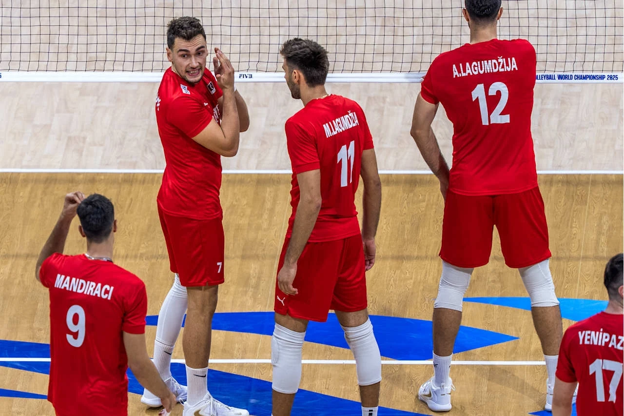 Grupa siatkarzy ubranych w czerwone stroje sportowe stoi na boisku, zawodnicy przygotowują się do akcji lub omawiają strategię, w tle siatka do siatkówki oraz fragment hali sportowej.