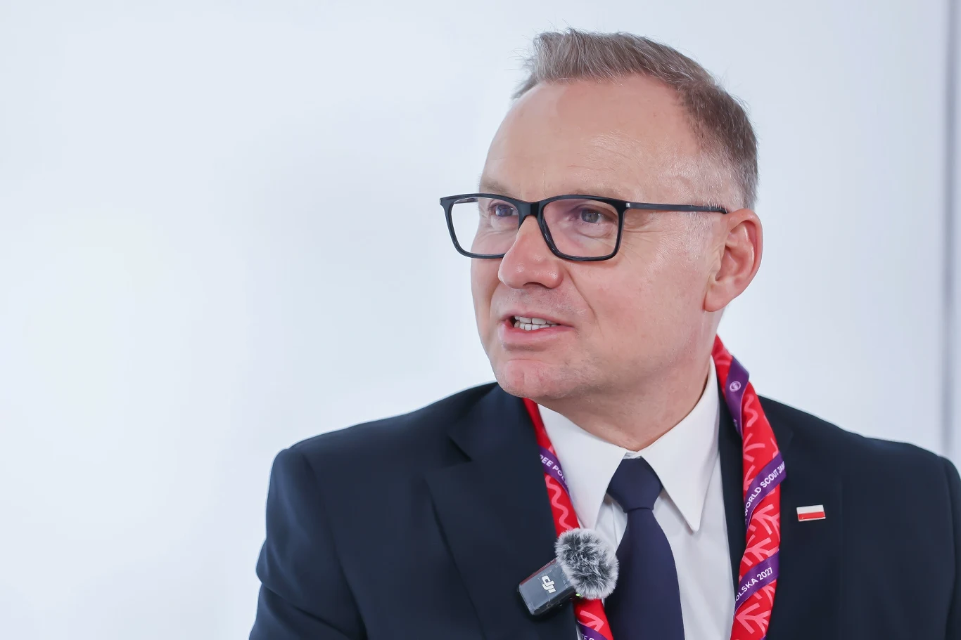Andrzej Duda reaguje na słowa Donalda Trumpa ws. rosyjskich dronów Mężczyzna w okularach w garniturze i krawacie, z przypiętym mikrofonem oraz wstążką o czerwonym kolorze, siedzi na tle jasnego, neutralnego tła.