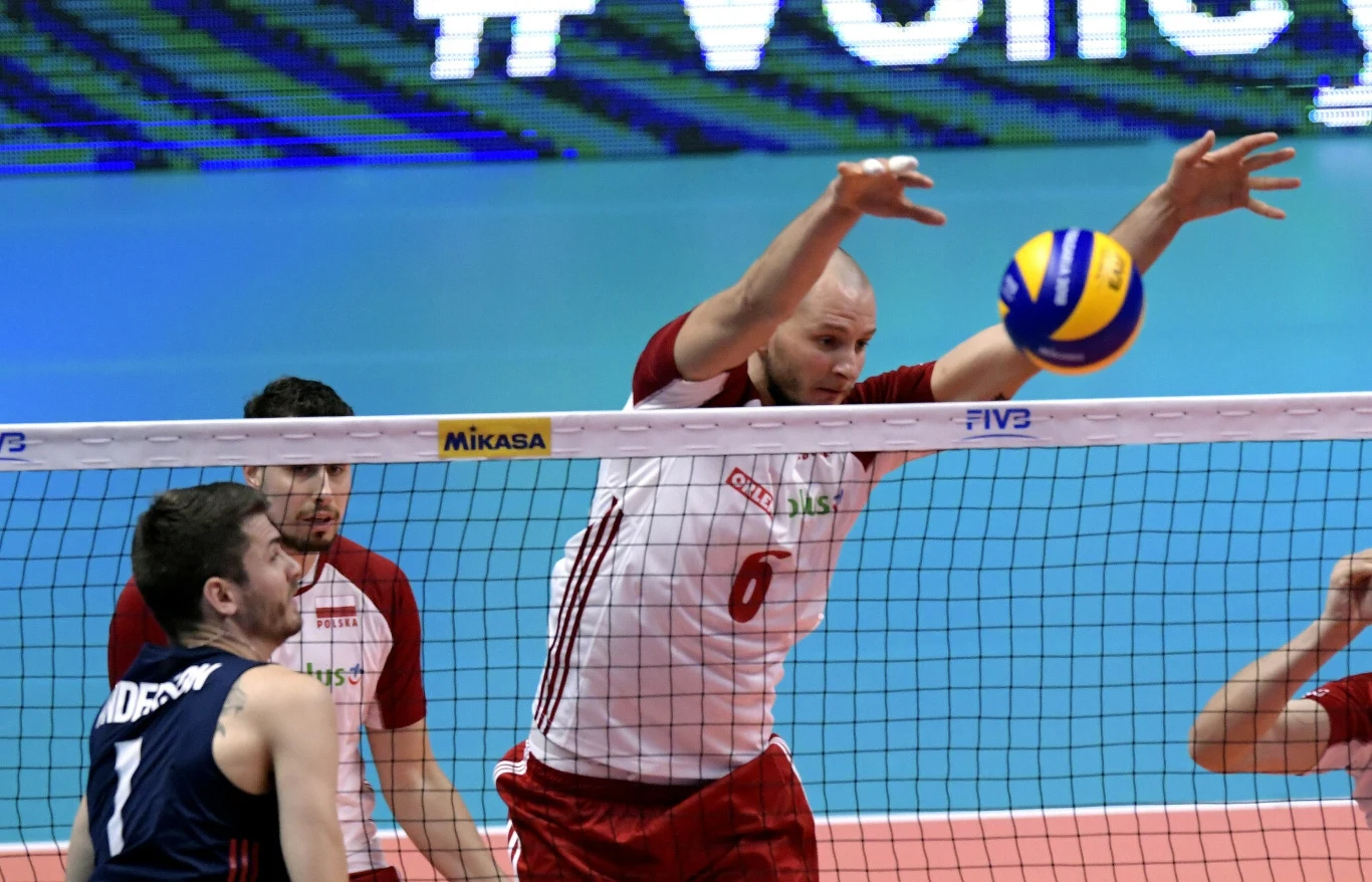 Dwóch siatkarzy w biało-czerwonych strojach walczy o piłkę tuż nad siatką podczas meczu, po lewej stronie widoczny jest przeciwnik w granatowym stroju próbujący zablokować atak.