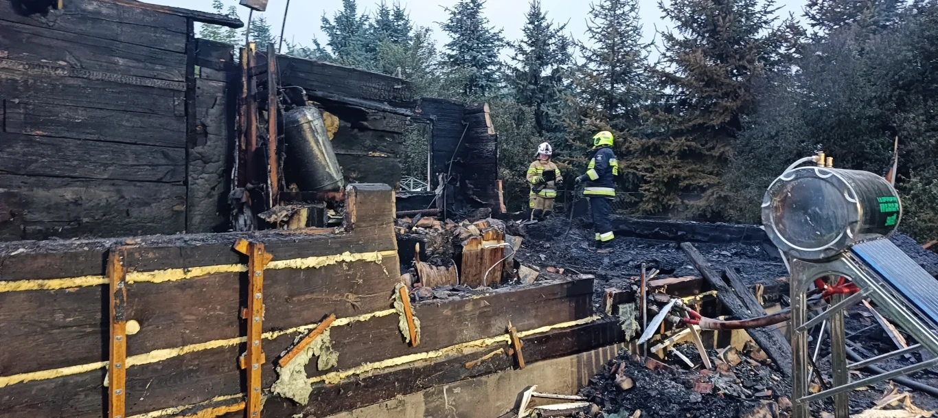 Pożar domu we wsi Kuzawka w gminie Terespol Dwie osoby w strojach strażackich stojące wśród zgliszczy spalonego drewnianego budynku otoczonego drzewami, widoczne szczątki konstrukcji i spalone przedmioty.