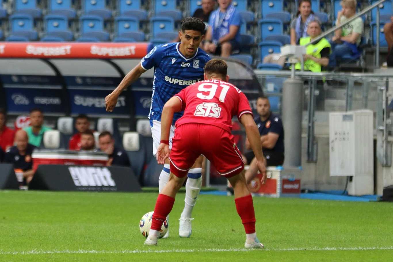 Dwóch piłkarzy w trakcie akcji na boisku, zawodnik w niebieskiej koszulce drybluje piłkę, podczas gdy drugi w czerwonej koszulce próbuje go zatrzymać, w tle widoczne trybuny z kibicami i ławka rezerwowych.