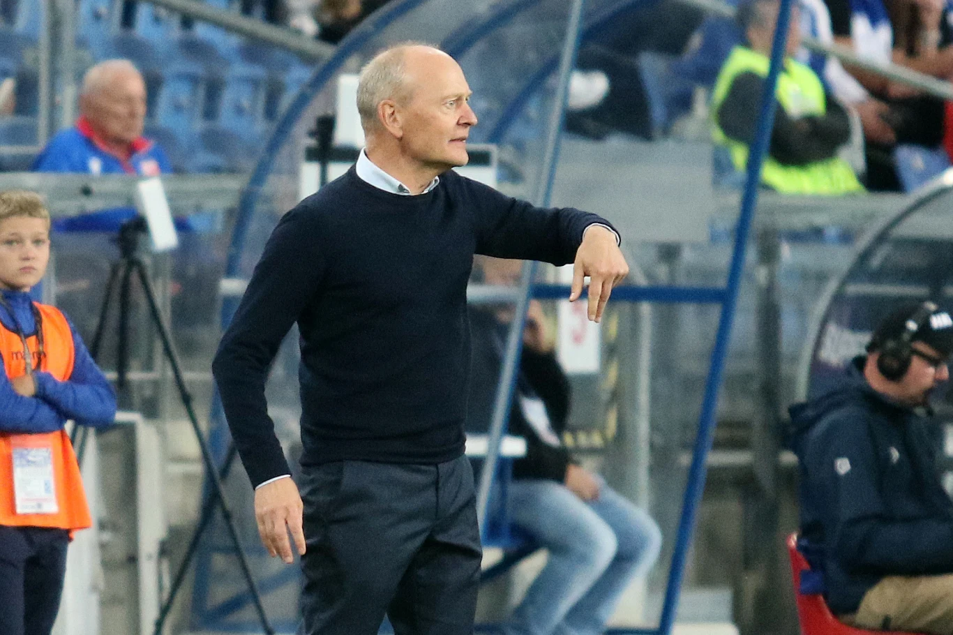 Trener w ciemnym swetrze gestykuluje na linii bocznej stadionu podczas meczu piłkarskiego, w tle widzowie oraz asystent z kamerą.