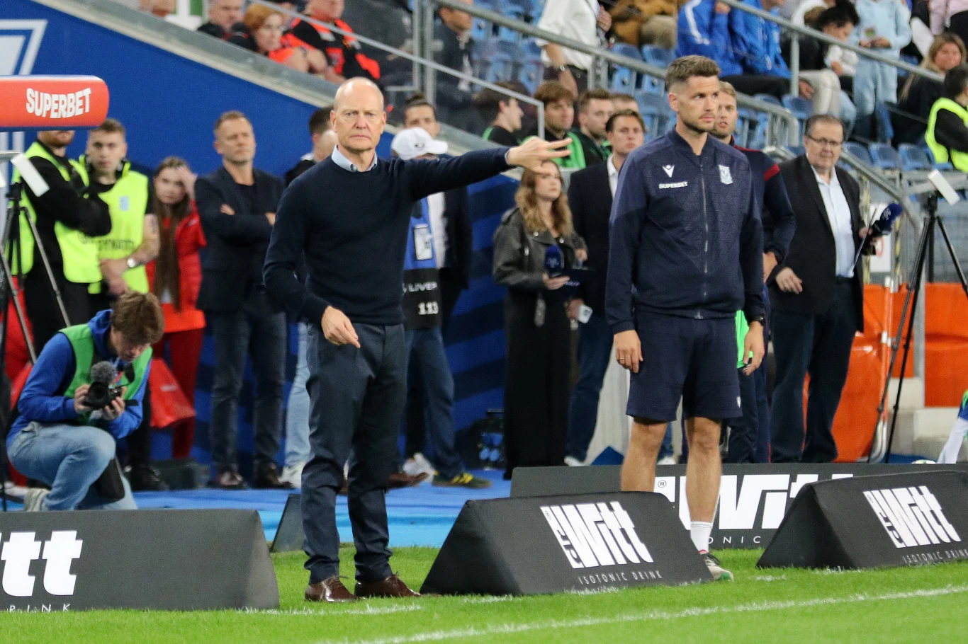 Trener oraz członek sztabu szkoleniowego stoją przy bocznej linii boiska, wskazując i wydając polecenia, w tle widoczni są kibice, obsługa techniczna oraz elementy stadionu.