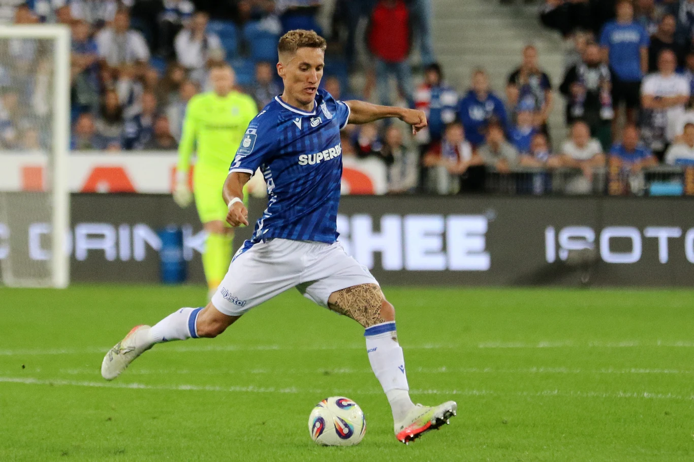 Joel Pereira, Lech Poznań Piłkarz w niebiesko-białym stroju wykonuje dynamiczny strzał na boisku podczas meczu piłkarskiego, w tle widoczna publiczność na trybunach oraz zawodnicy obu drużyn.