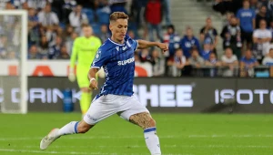 Piłkarz w niebiesko-białym stroju wykonuje dynamiczny strzał na boisku podczas meczu piłkarskiego, w tle widoczna publiczność na trybunach oraz zawodnicy obu drużyn.