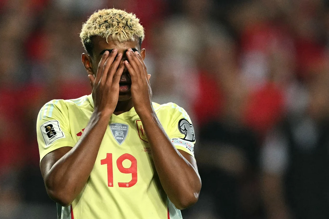 Lamine Yamal Piłkarz w jasnej koszulce z numerem 19 i rękami zakrywającymi twarz podczas meczu, w tle niewyraźni kibice na trybunach.