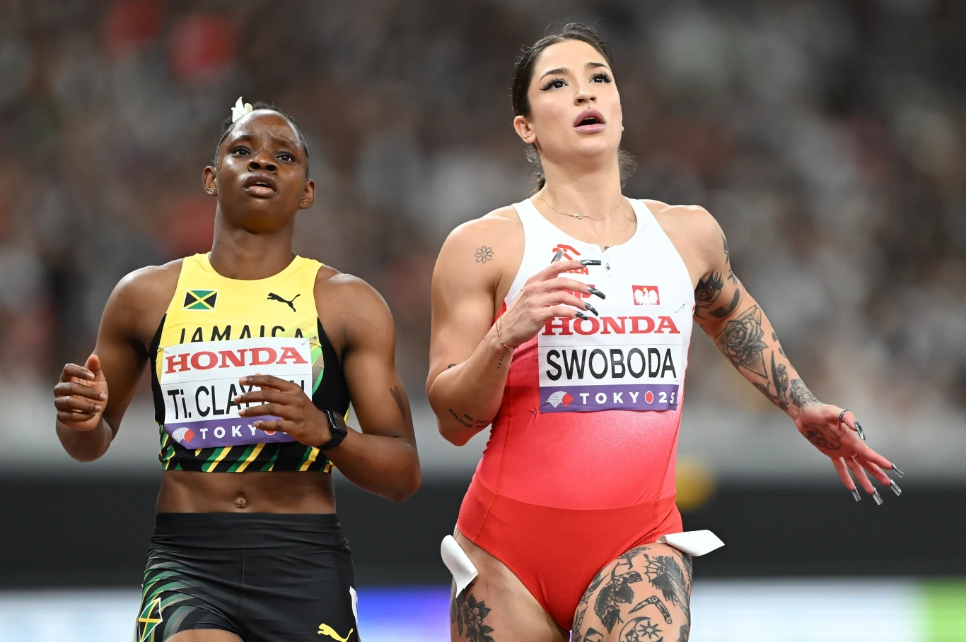 Ewa Swoboda i Tina Clayton Dwóch zawodniczek biegnących na stadionie lekkoatletycznym podczas zawodów, jedna reprezentuje Jamajkę, druga Polskę, obie w sportowych strojach z numerami startowymi i sponsorami, w tle widownia.