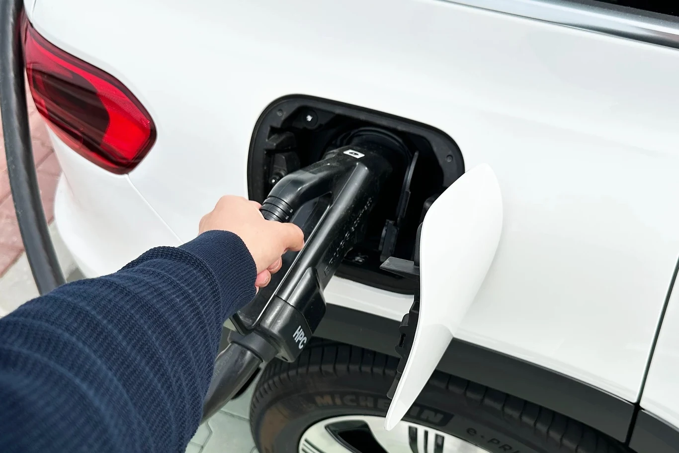 Kable do ładowania samochodów elektrycznych coraz częściej padają łupem złodziei Ręka trzymająca pistolet do tankowania paliwa, wlew umieszczony w białym samochodzie, widoczna tylna część pojazdu i opona.