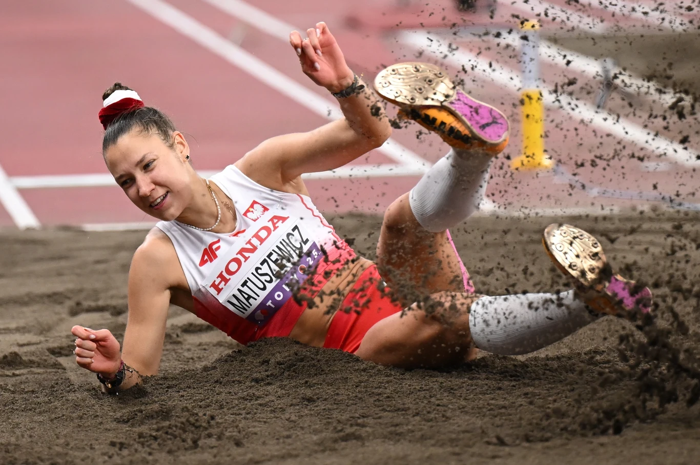 Lekkoatletka w sportowym stroju wykonuje skok w dal, lądując w piasku z dynamicznym wyrazem twarzy, wokół widoczny rozprysk ziemi, a na tle widać fragment bieżni stadionowej.