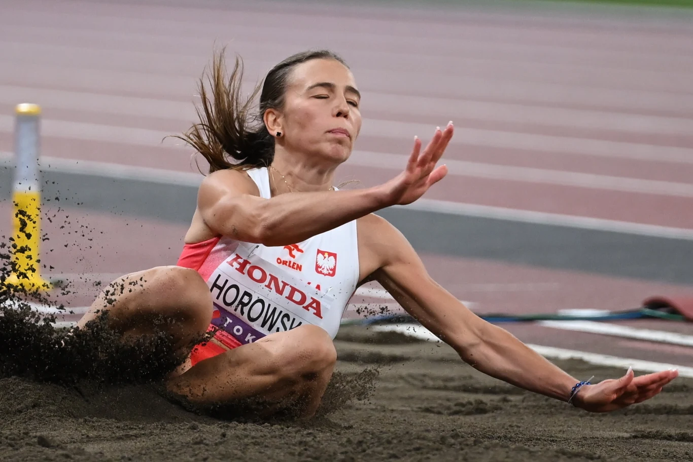 Zawodniczka lekkoatletyczna wykonuje skok w dal, lądując w piaskownicy, z zamkniętymi oczami i skupioną twarzą. Na jej koszulce widnieje napis z nazwą sponsora oraz narodowe godło Polski.