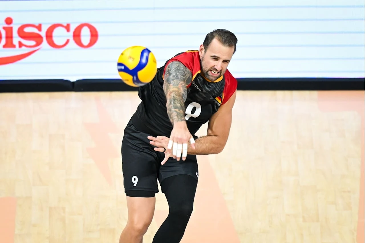 Siatkarz z widocznym tatuażem na ramieniu odbijający piłkę podczas meczu na hali sportowej; dynamiczny moment akcji sportowej.