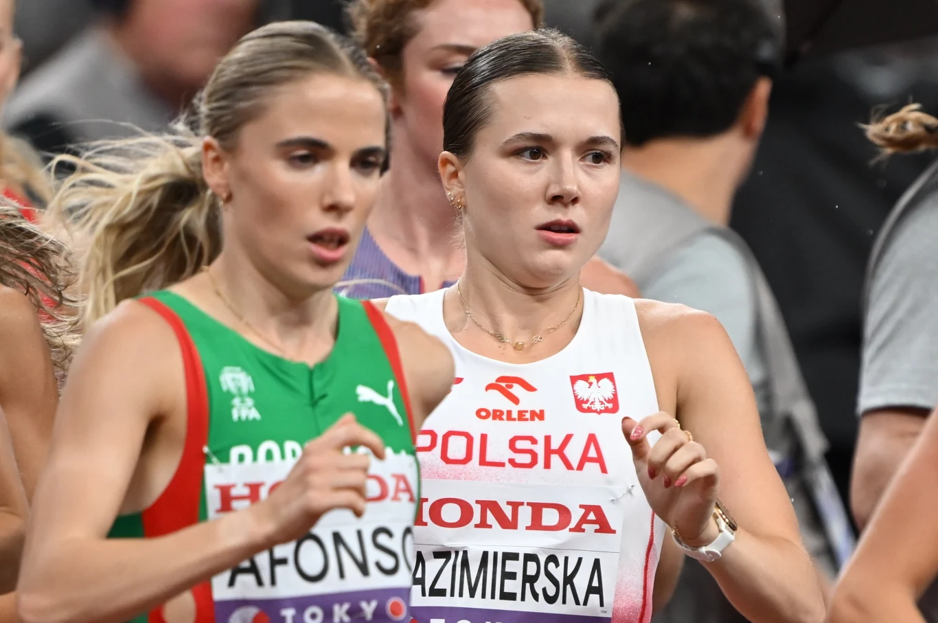 Dwie biegaczki rywalizują podczas wyścigu, jedna z Polski w białym stroju z godłem narodowym, druga z Portugalii w zielono-czerwonym stroju, obie skupione i intensywnie biegnące w tłumie innych zawodniczek.