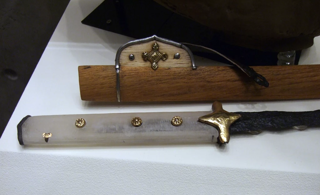Stara japońska katana z bogato zdobioną rękojeścią oraz oprawą zawierającą elementy metalowe i drewniane, leżąca na białym podłożu wystawowym.