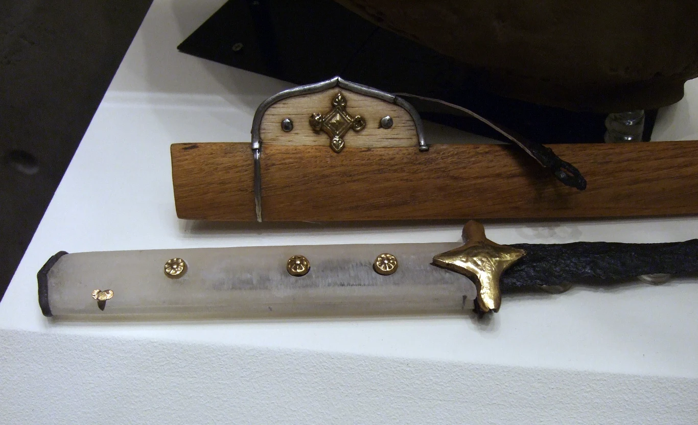 Stara japońska katana z bogato zdobioną rękojeścią oraz oprawą zawierającą elementy metalowe i drewniane, leżąca na białym podłożu wystawowym.