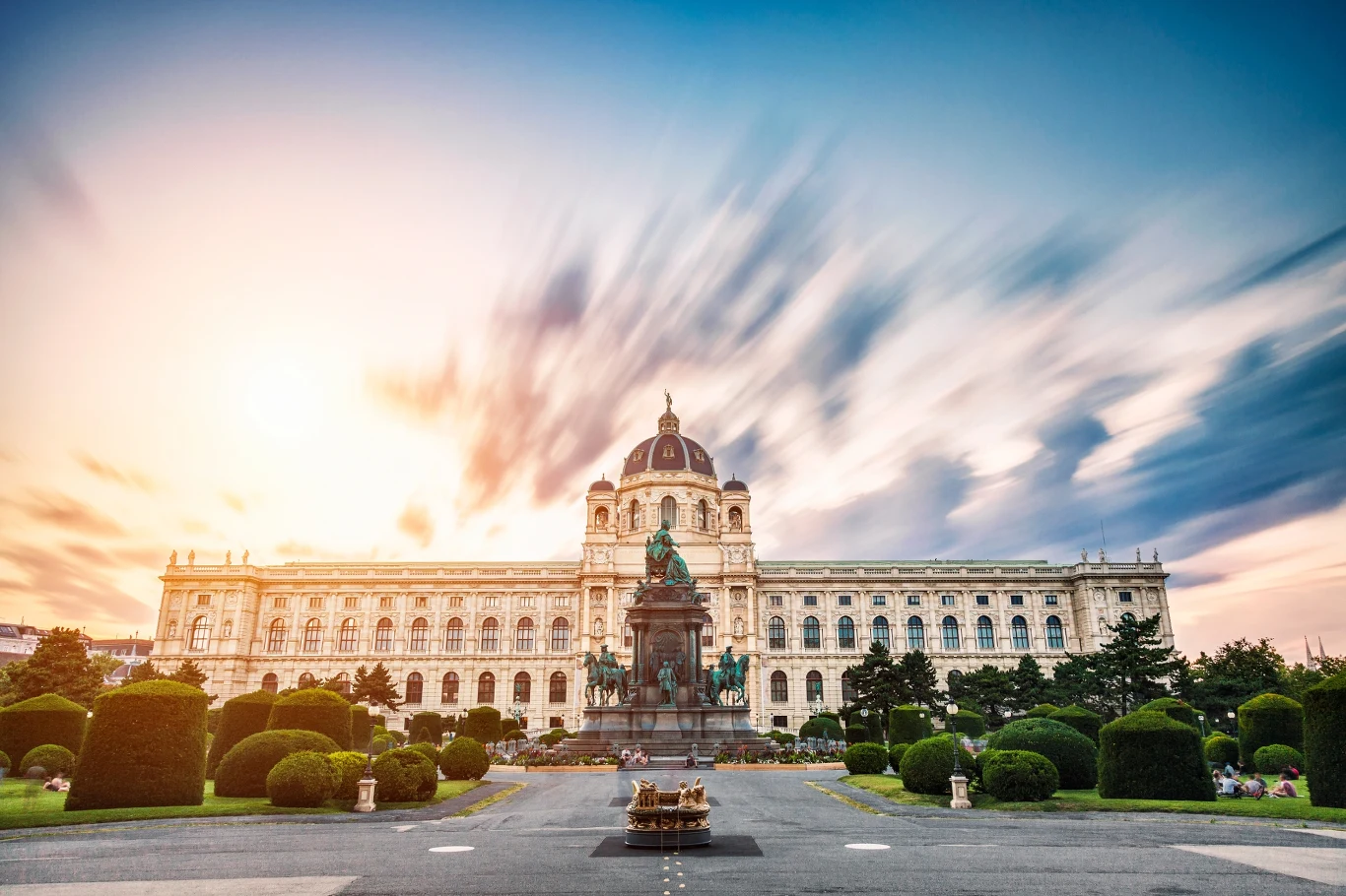 Monumentalny gmach muzeum z dużą kopułą pośrodku, otoczony starannie przyciętymi krzewami i alejkami, na pierwszym planie znajduje się pomnik konny ustawiony na wysokim cokole, nad całością rozpościera się kolorowe, dynamiczne niebo.