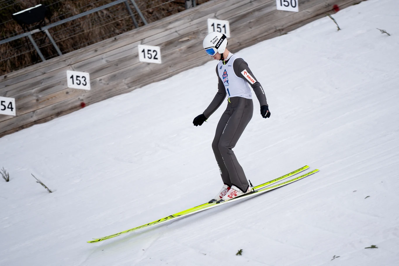 Kacper Tomasiak Zawodnik skaczący na nartach podczas konkursu skoków narciarskich w locie tuż po lądowaniu na śniegu, w tle numery oznaczające odległości na skoczni
