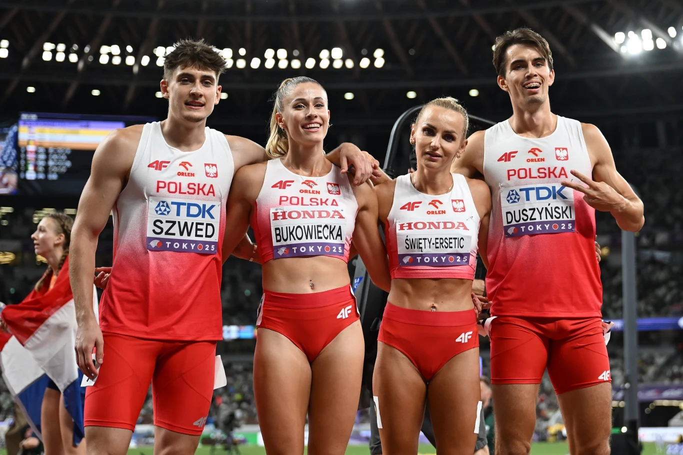 Czwórka polskich lekkoatletów w stroju reprezentacyjnym po zakończonym biegu na stadionie lekkoatletycznym, uśmiechnięci, obejmują się wzajemnie, w tle trybuny i kibice.