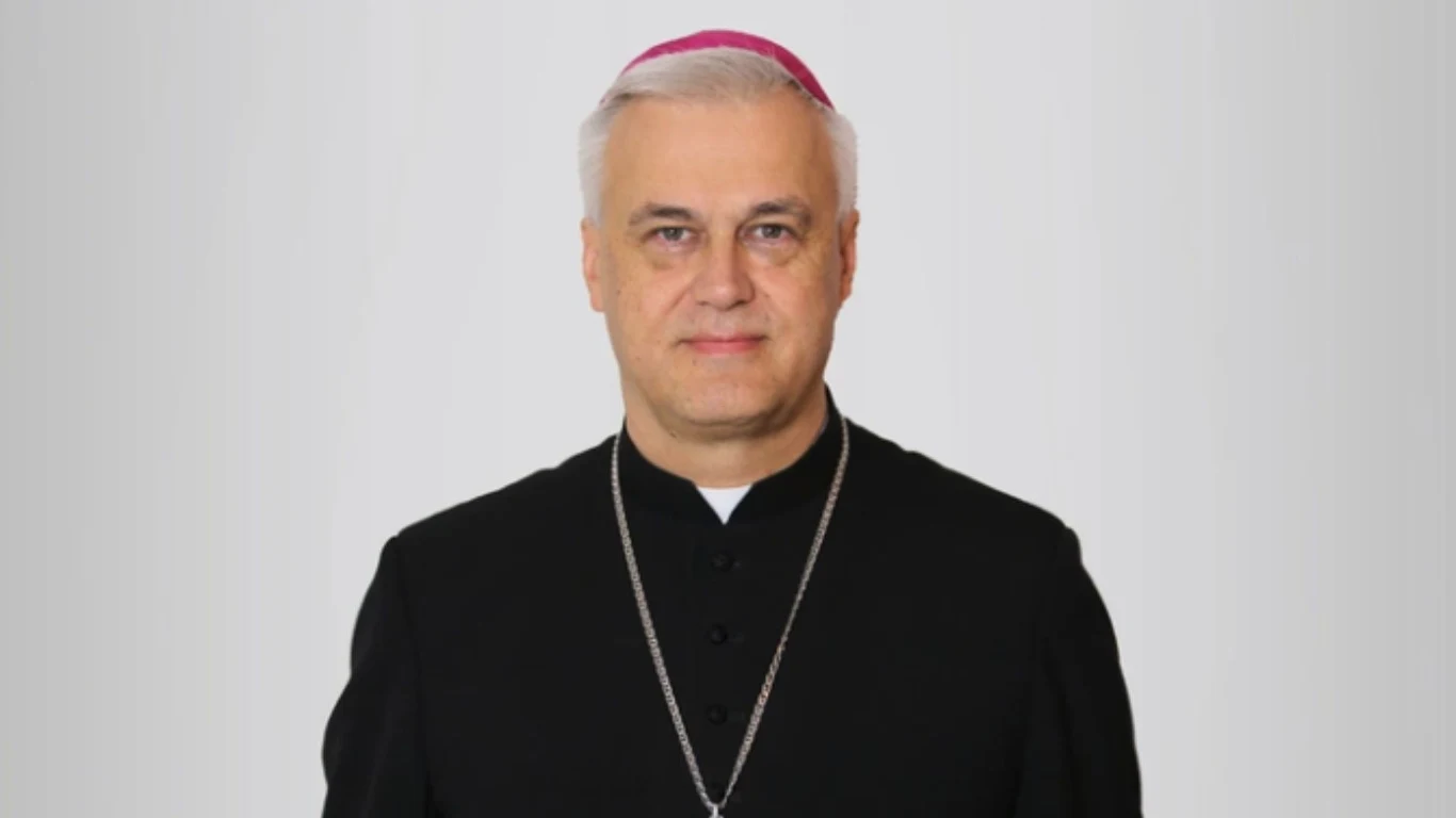 Bp Wojciech Skibicki Mężczyzna w średnim wieku z siwymi włosami, ubrany w czarną sutannę z białą koloratką oraz fioletową piuską na głowie, na szyi zawieszony srebrny łańcuszek.