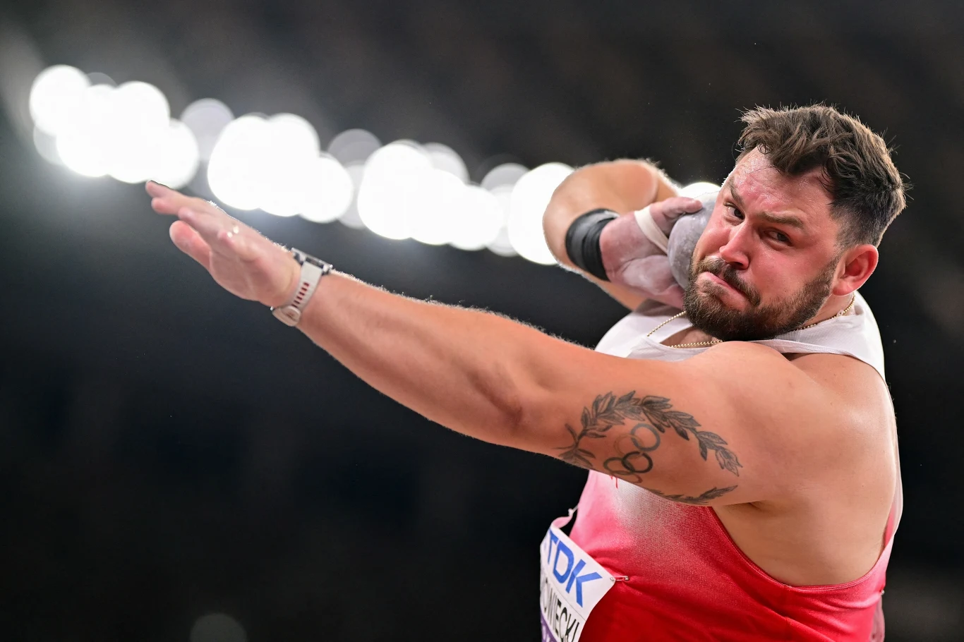 Lekkoatleta w trakcie oddawania rzutu kulą, skupiony, ubrany w sportowy strój z odsłoniętym ramieniem, na którym widoczny jest tatuaż, dynamiczna poza, wyraziste oświetlenie stadionowe w tle.