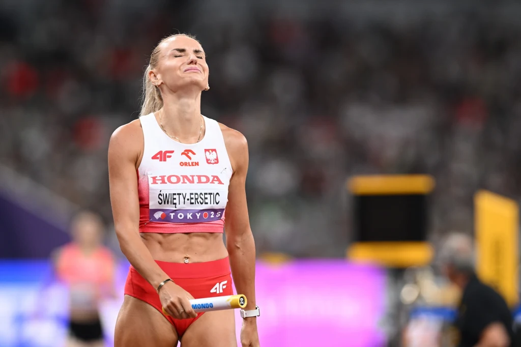 Sportsmenka w stroju startowym z wyrazem zmęczenia lub rozczarowania na twarzy, trzymająca pałeczkę sztafetową na stadionie lekkoatletycznym podczas zawodów.