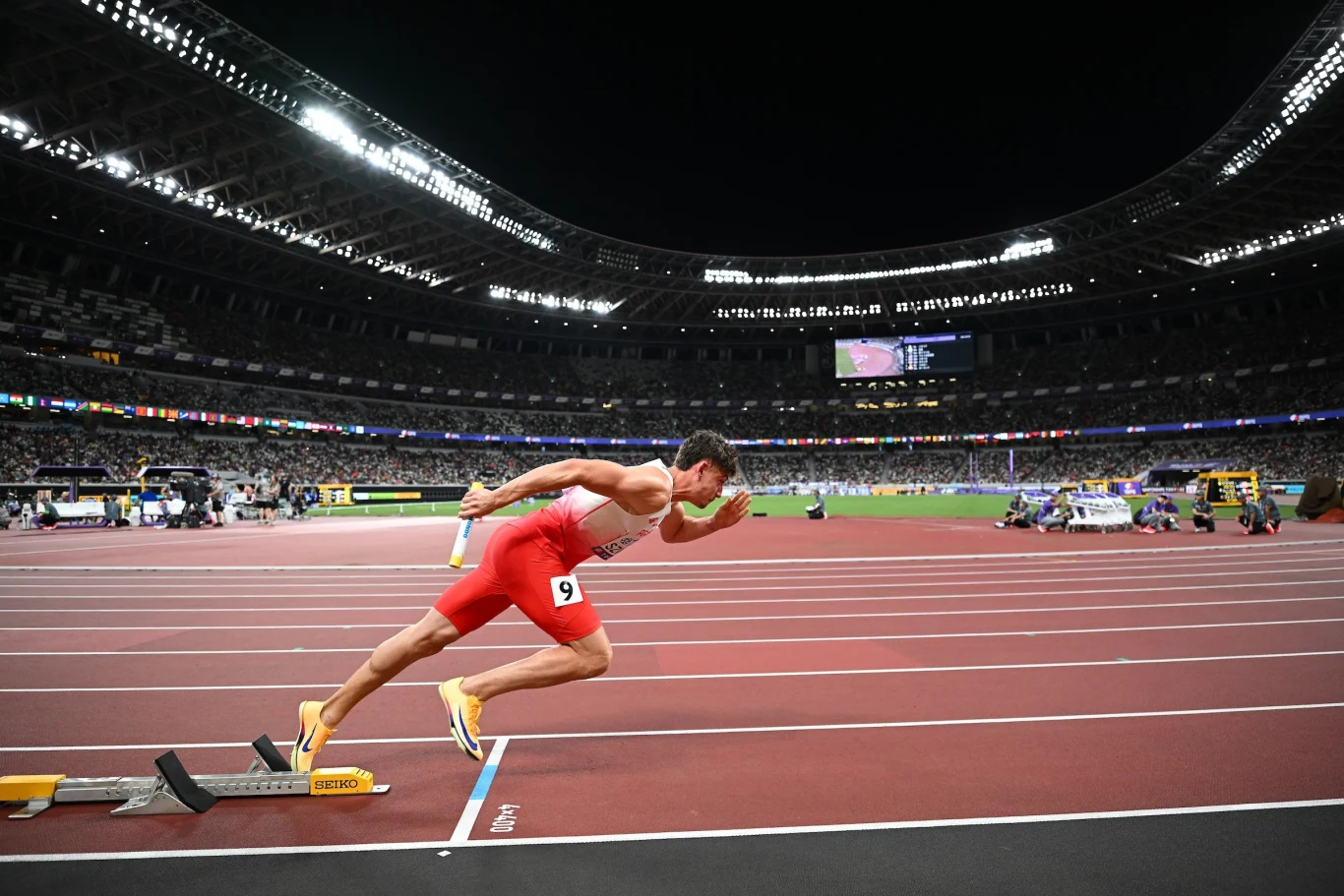 Lekkoatleta w czerwonym stroju startuje ze stanowiska na stadionie lekkoatletycznym w nocy, w tle widoczne trybuny pełne widzów oraz nowoczesne oświetlenie stadionu.