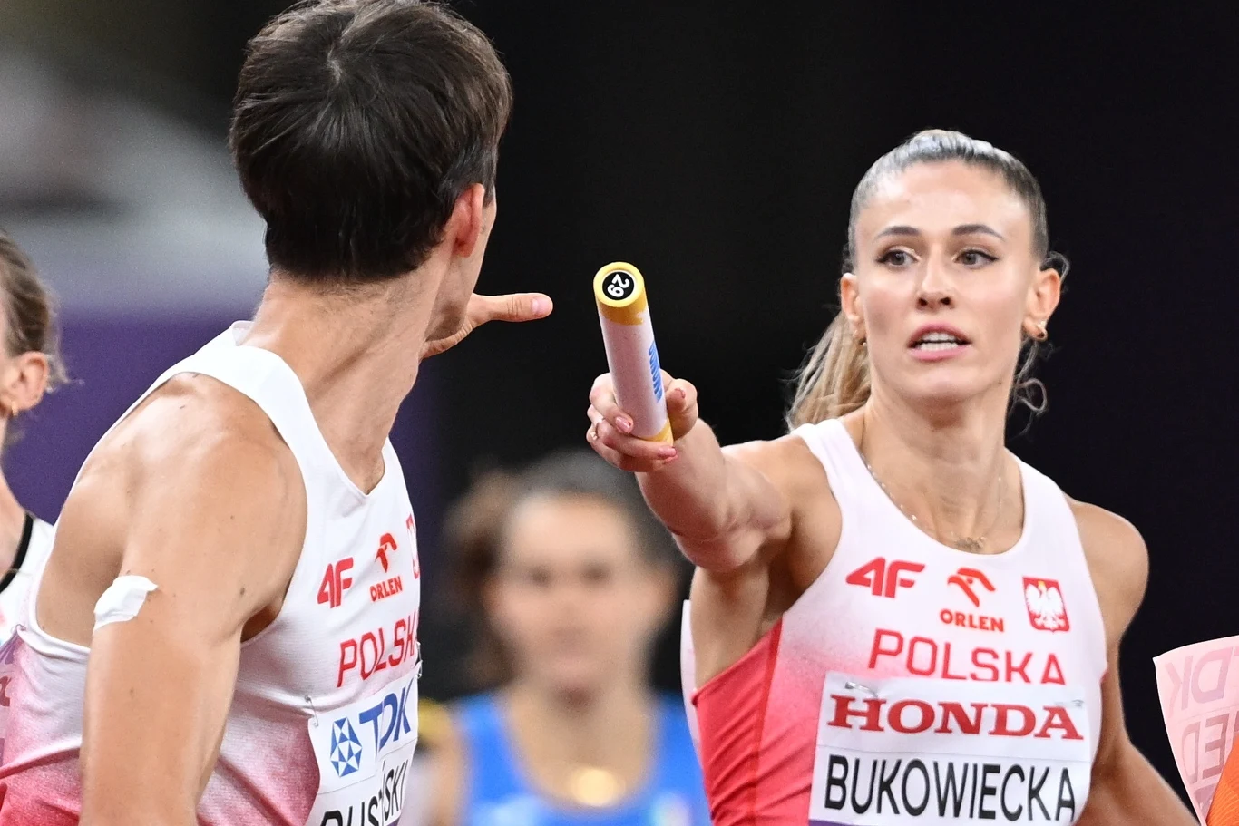 Zawodniczka polskiej sztafety przekazuje pałeczkę koledze z drużyny podczas biegu sztafetowego, oboje skupieni na zadaniu, w tle rozmyte postacie innych zawodników