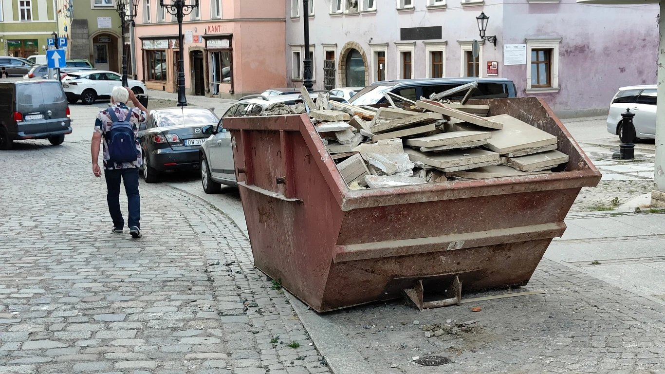 Skutki powodzi z 2024 roku nadal widać na ulicach Kłodzka i nie tylko Kontener wypełniony gruzem i odpadami budowlanymi ustawiony na brukowanej ulicy, w tle zaparkowane samochody oraz przechodzień idący chodnikiem obok zabytkowych kamienic.