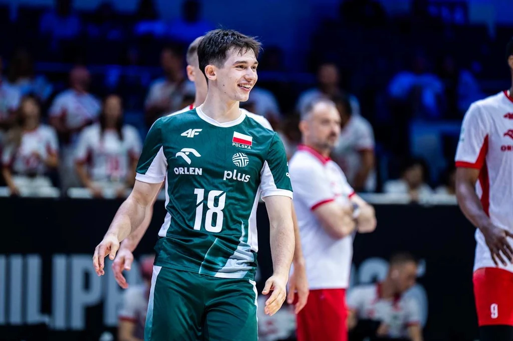 Maksymilian Granieczny w meczu z Rumunią Młody siatkarz ubrany w zielony strój reprezentacji Polski stoi na boisku podczas meczu, uśmiecha się, wokół widać innych zawodników oraz trenerów w tle.
