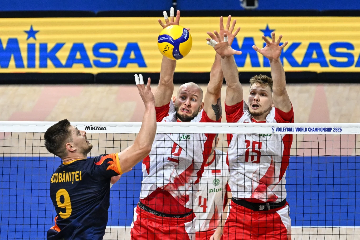 Jakub Kochanowski (z prawej) i Bartosz Kurek Dwóch siatkarzy w biało-czerwonych strojach blokuje piłkę nad siatką, podczas gdy przeciwnik z drużyny w granatowo-pomarańczowym stroju próbuje zaatakować. W tle widoczna reklama firmy Mikasa, a na boisku wyraźna linia oddzielająca pole gry.