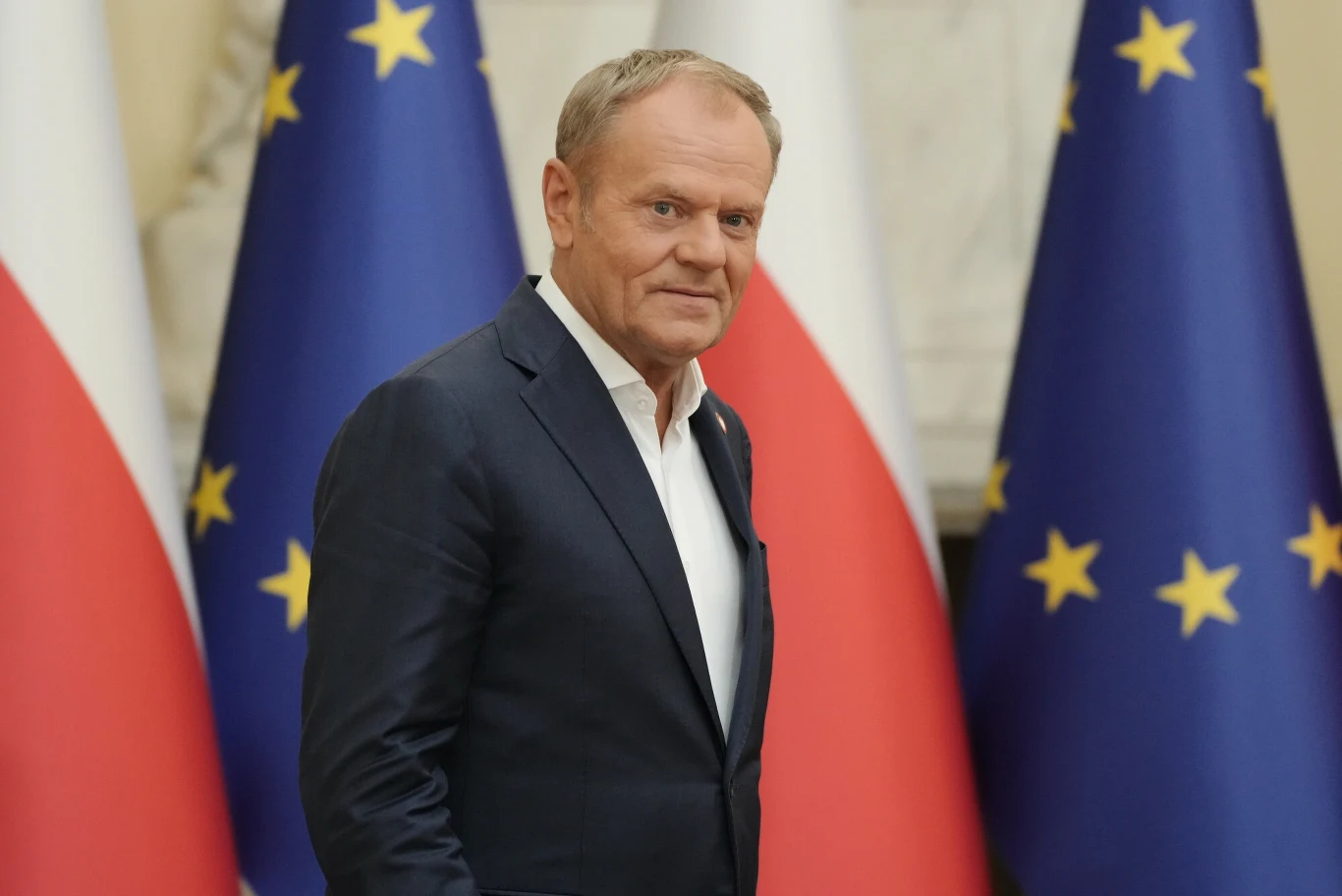 Premier Donald Tusk Mężczyzna w garniturze stoi na tle flag Polski i Unii Europejskiej, patrzy lekko w bok, w tle widoczne są jasne ściany.