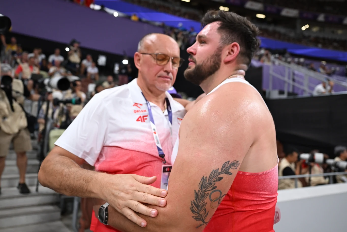 Sportowiec w stroju startowym i jego trener w biało-czerwonej koszulce obejmują się na stadionie lekkoatletycznym, obaj wyrażają silne emocje po zakończonej rywalizacji.