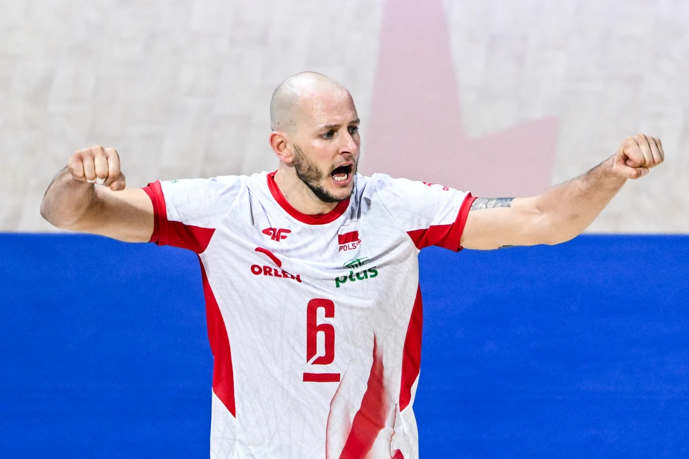 Bartosz Kurek Siatkarz w białym stroju z czerwonymi akcentami i numerem 6 na koszulce, unosi obie ręce w geście triumfu lub energii, z wyrazem determinacji na twarzy, na tle niebieskiego boiska.