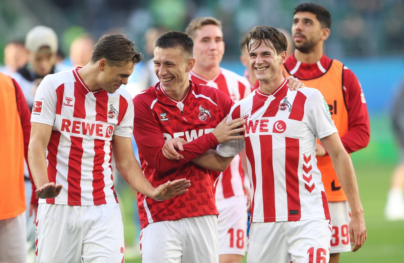 Jakub Kamiński (z prawej) został bohaterem 1. FC Koeln podczas starcia z VfL Wolfsburg Kilku piłkarzy drużyny 1. FC Köln ubranych w charakterystyczne biało-czerwone stroje z widocznym logo sponsora, uśmiecha się i cieszy się po zakończonym meczu na tle murawy stadionu.