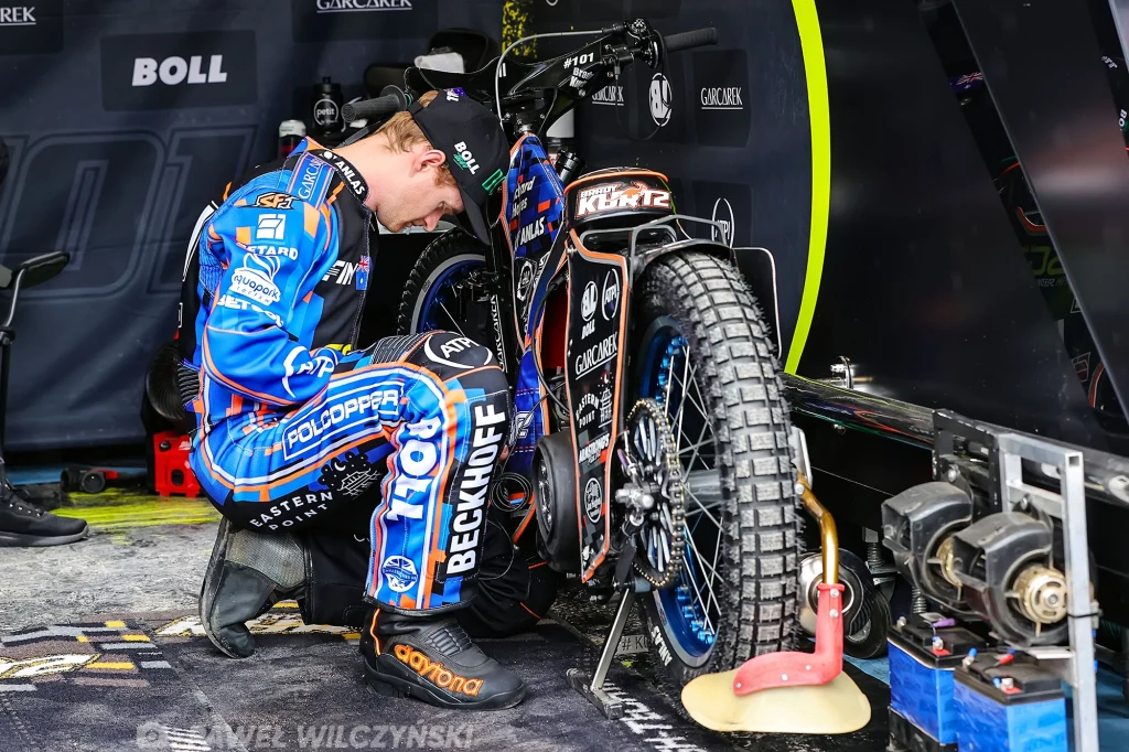 Zawodnik ubrany w profesjonalny kombinezon sportowy pochyla się przy motocyklu żużlowym, koncentrując się na elementach technicznych maszyny w przygotowaniu do zawodów